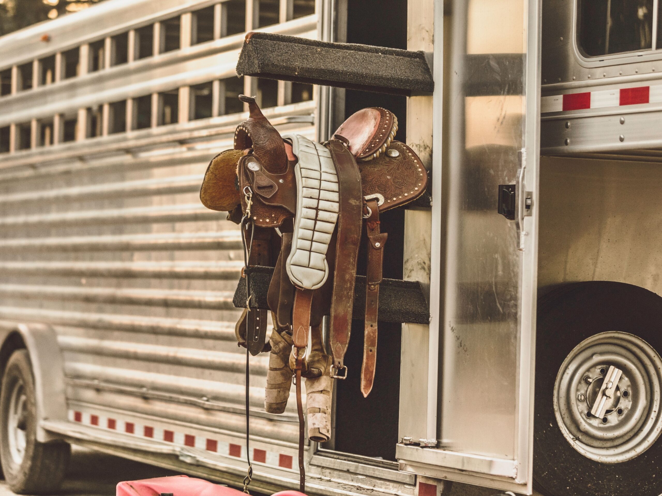 A detailed shot of western tack, including a leather saddle and gear, stored on an external rack of a silver horse trailer.