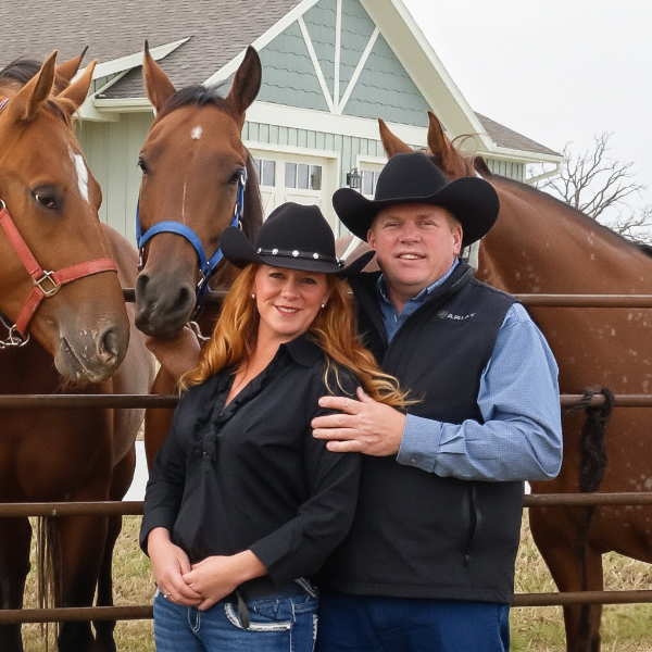 A couple wearing cowboy hats standing by a fence with several horses behind them.