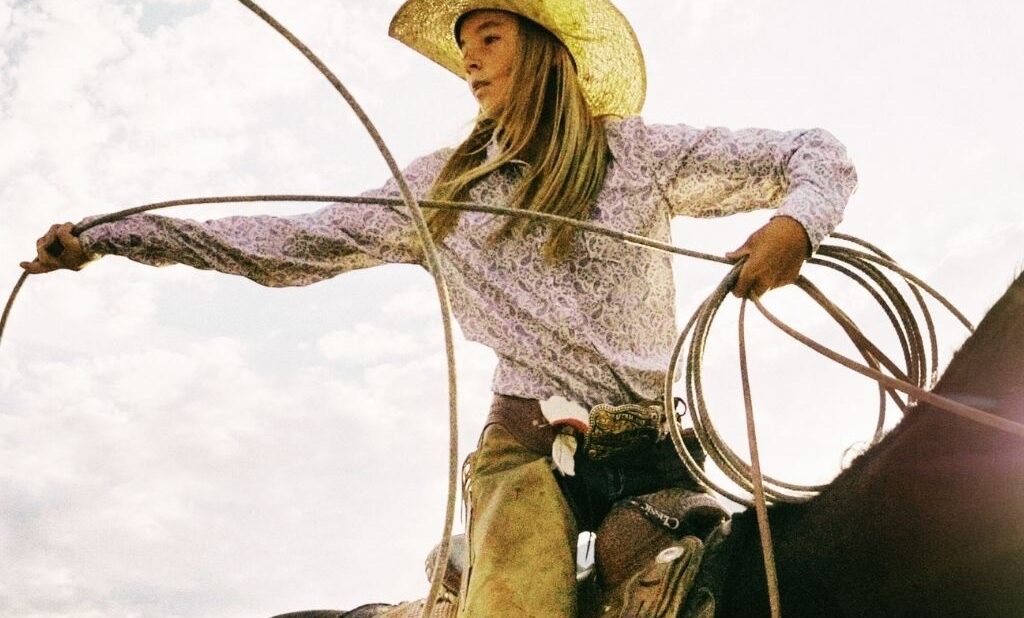 Close-up of a female rider in traditional western gear, including a wide-brimmed hat and leather chaps, demonstrating how to hold a lasso for roping.