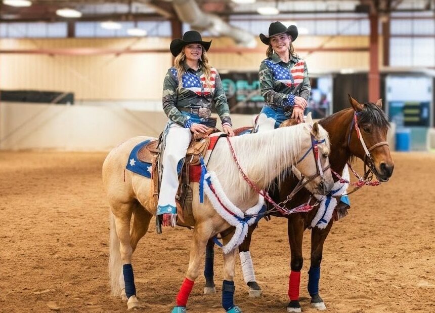 A wide shot of an indoor riding arena where two equestrian performers stand stationary on their horses. Both the riders and horses are fully outfitted in coordinated red, white, and blue gear for a themed horse show or training session.