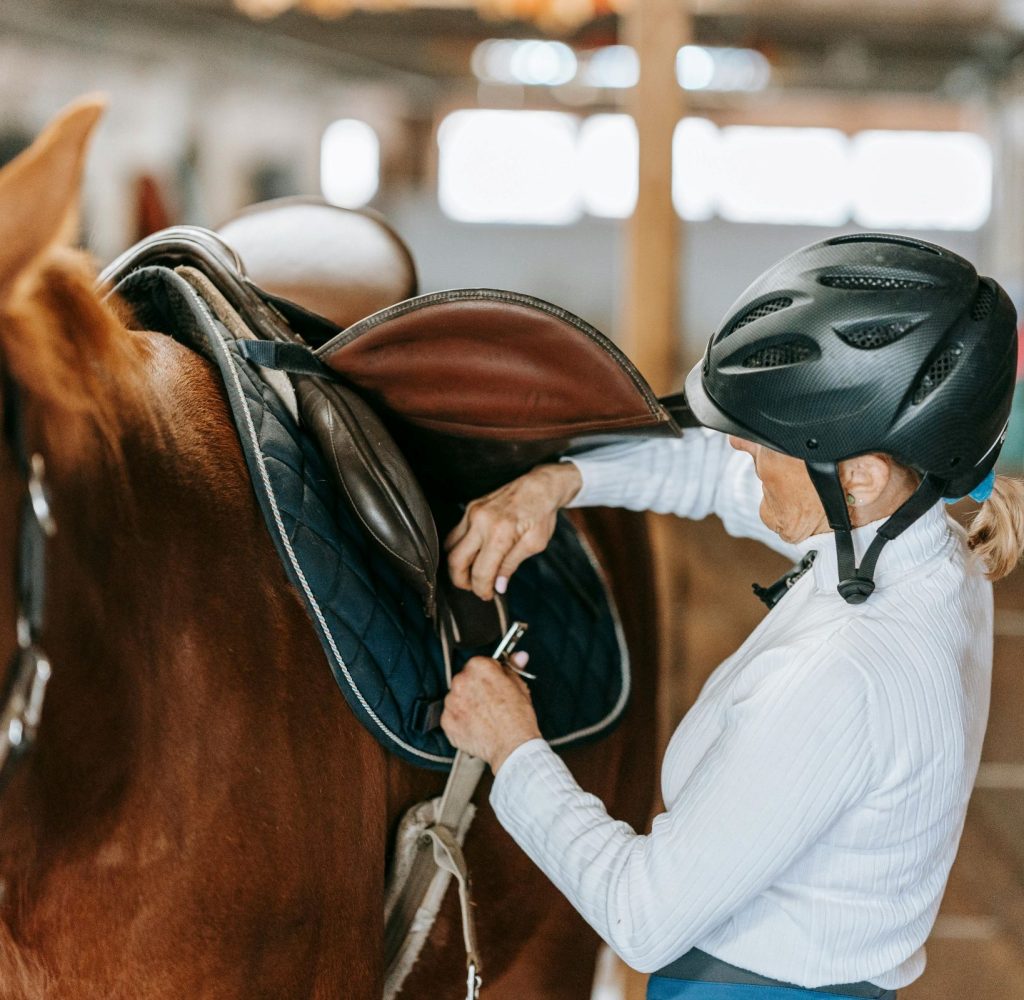 A person wearing a black riding helmet and a white long-sleeved shirt is shown from the side, focused on securing the girth of a brown leather saddle on a chestnut horse. The horse, which has a white marking on its face, stands patiently in a brightly lit barn aisle with wooden stalls visible in the background.